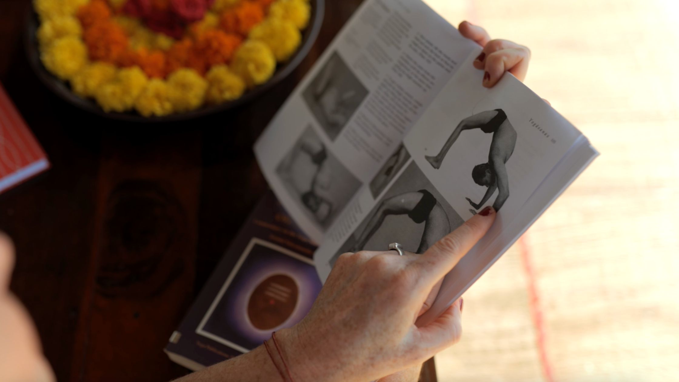 Close-up of a hand pointing at a traditional yoga book showing a black and white image of an asana, with marigold flowers in the background